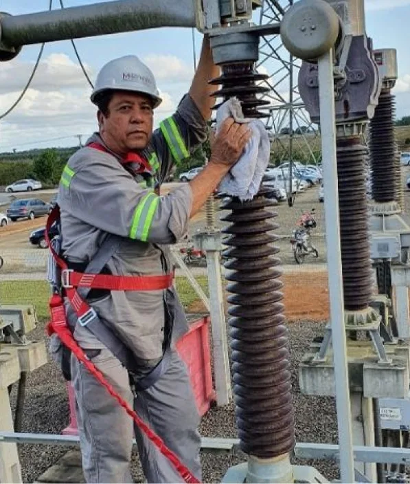 Técnico de capacete com cinto de segurança limpando o isolador de um equipamento de subestação